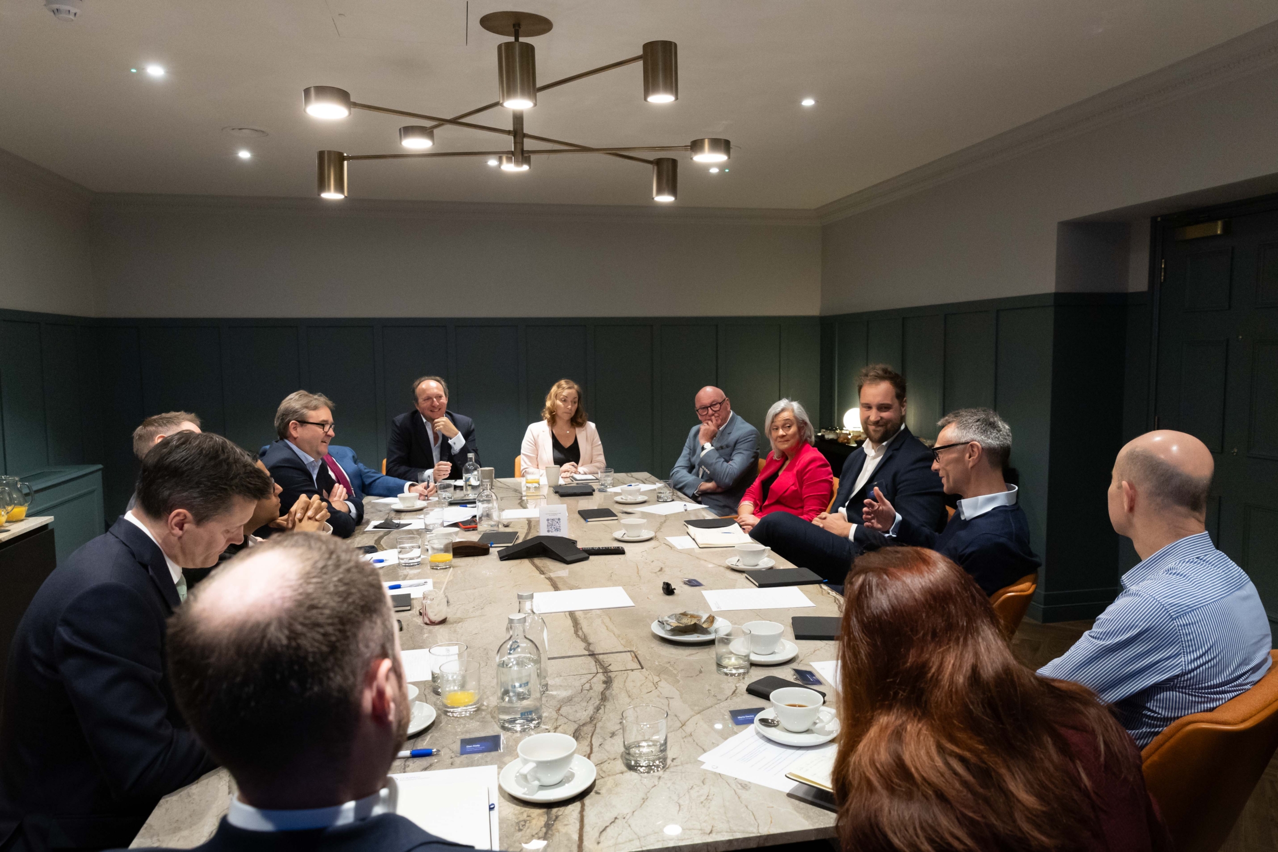 Compass Carter Osborne - Alt Text: A group of business professionals sitting around a large, rectangular conference table, engaged in a meeting. The room is well-lit with modern overhead lighting, and the table is set with notebooks, documents, glasses of water, and coffee cups. The attendees, both men and women, are dressed in business attire, and the mood appears to be focused and collaborative.